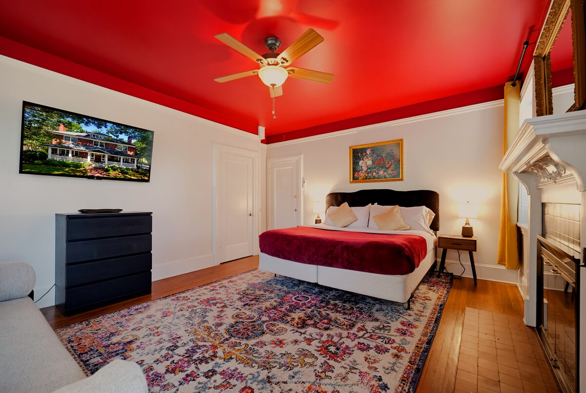 A wide-angle view of a bedroom with a red ceiling, a large bed, a TV mounted on the wall, a fireplace, and a patterned rug.