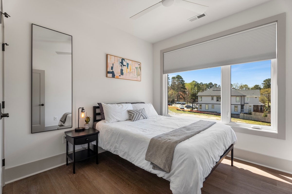 A neatly made bed with white bedding and a gray throw blanket, a nightstand with a lamp, and a large mirror on the wall in a room with a ceiling fan and a window with blinds.