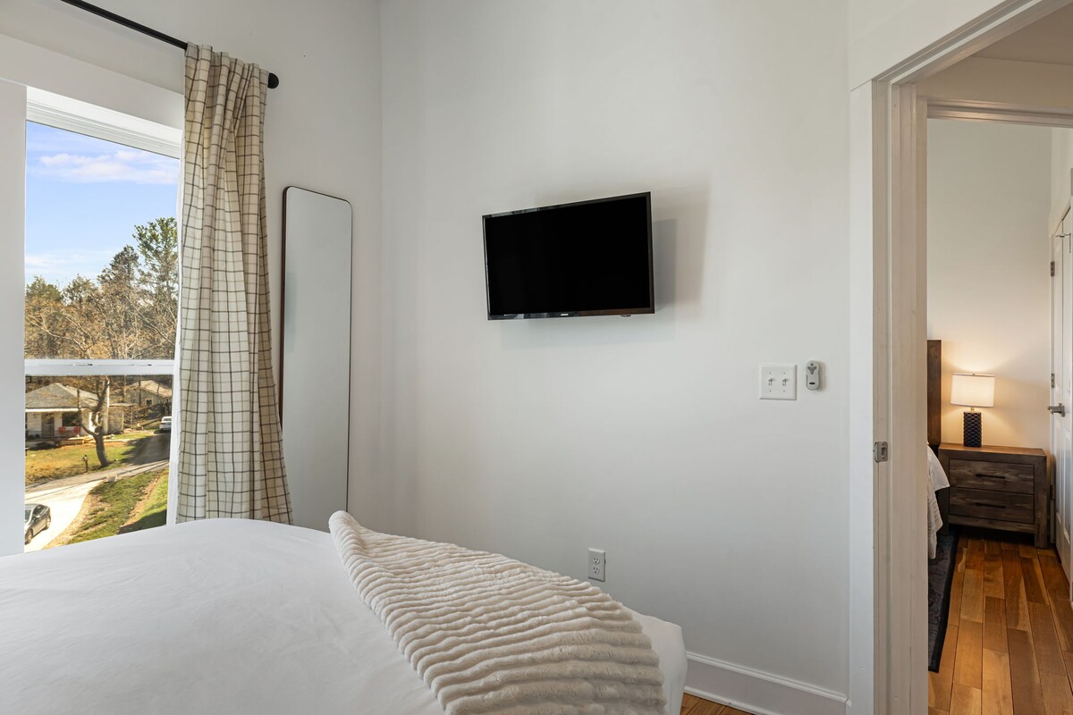 A wide view of a hotel room with a bed, window, TV, and doorway.