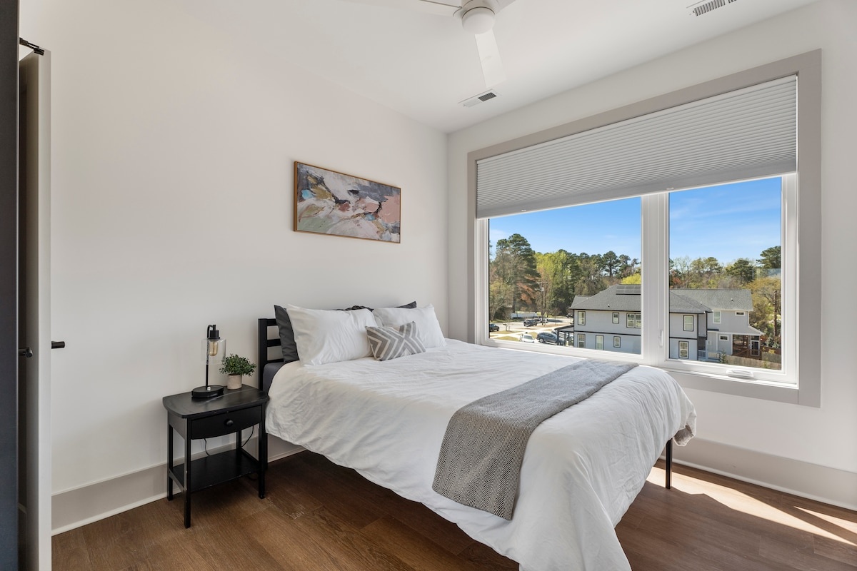 A neatly made bed with white linens and a gray throw blanket sits under a window with blinds, next to a small black nightstand with a lamp and plant.